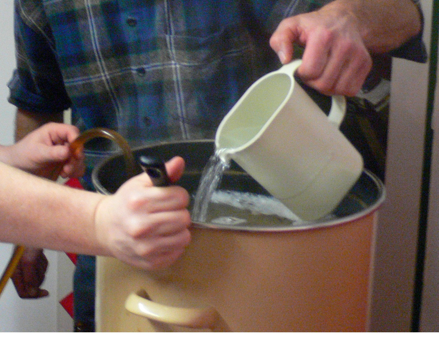 Im Vordergrund sieht man einen blas gelben Kochtopft. Danhinter steht eine Person, von der man nur einen Teil eines karrierten Hemdes sieht. Mit der einen Hand wird Wasser aus einem kleinen Gefäß in den Topf gegossen. Von der zweiten Person sieht man nur die Hände. In der einen wird ein Löffel gehalten, mit der anderen ein Schlauch. Beides ist im Topf. Die Flüssigkeit im Topf reicht fast bis zum Rand und eine leichte Schaumbildung ist erkennbar.
