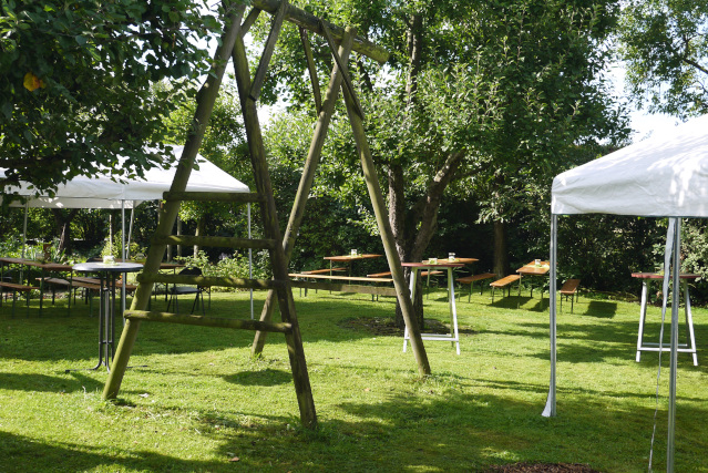 Grüne Wiese mit einem Schaukelgerüst (ohne Schaukel) umrahmt von herabhängenden Ästen. Linkst und rechts sind weiße Pavillione. Bierbänke und Stehtische sind im Garten verteilt.