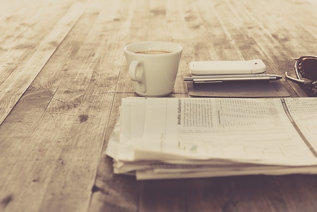 Alles ist in creme Farben gehalten. Eine Zeitung, eine Tsse, ein Handy und eine Brille liegen auf einem Holztisch. In der Tasse ist Kaffe und die Zeitung ist gefaltet, aber die Schrift nicht lesbar.